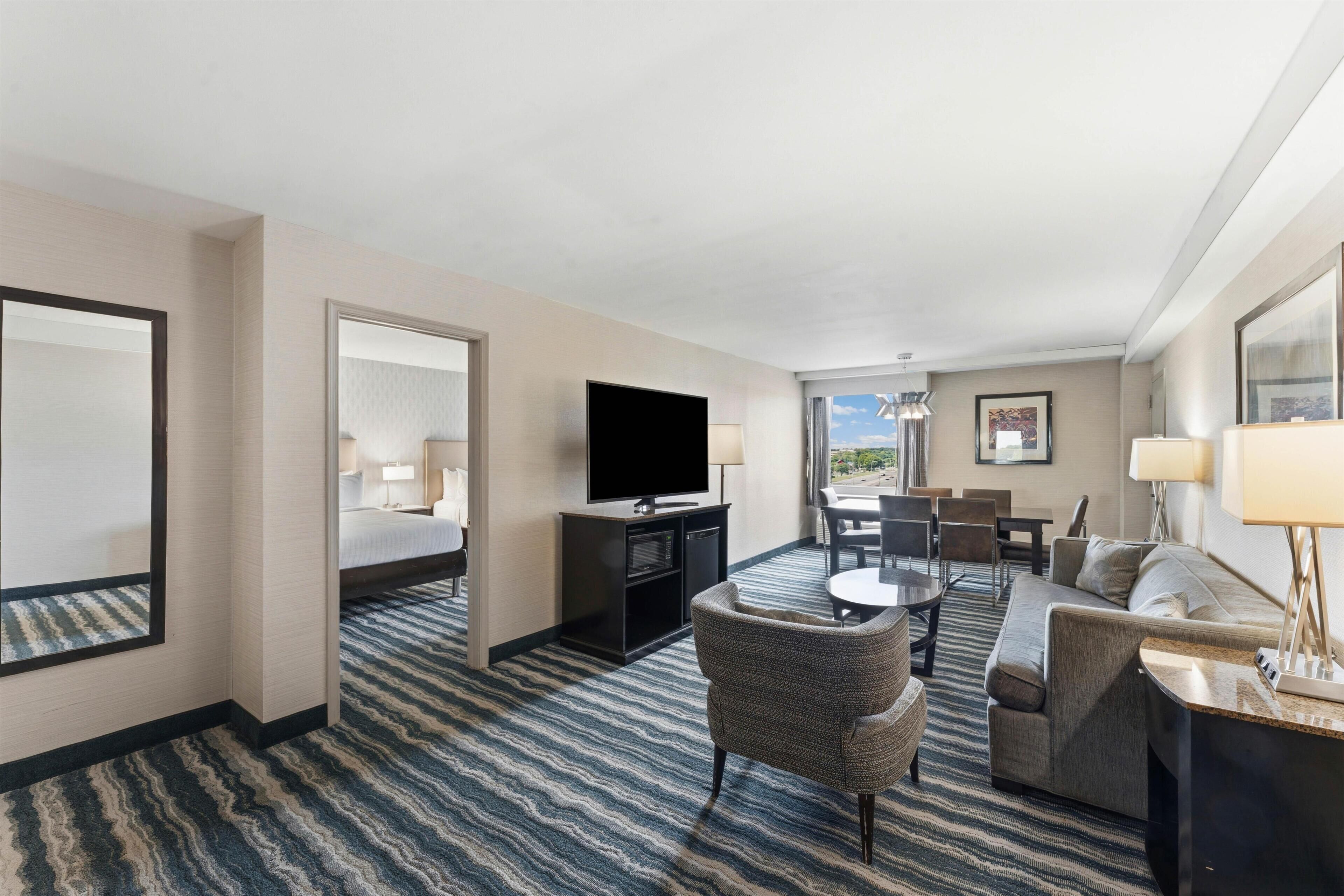 Quarto de hotel com cama, televisão, escrivaninha, sofá e área de refeições com cadeiras.