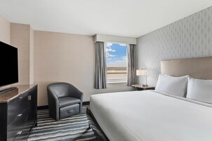 Una habitación de hotel con una cama, una silla, un televisor, una ventana con cortinas y una mesita de noche con lámpara.