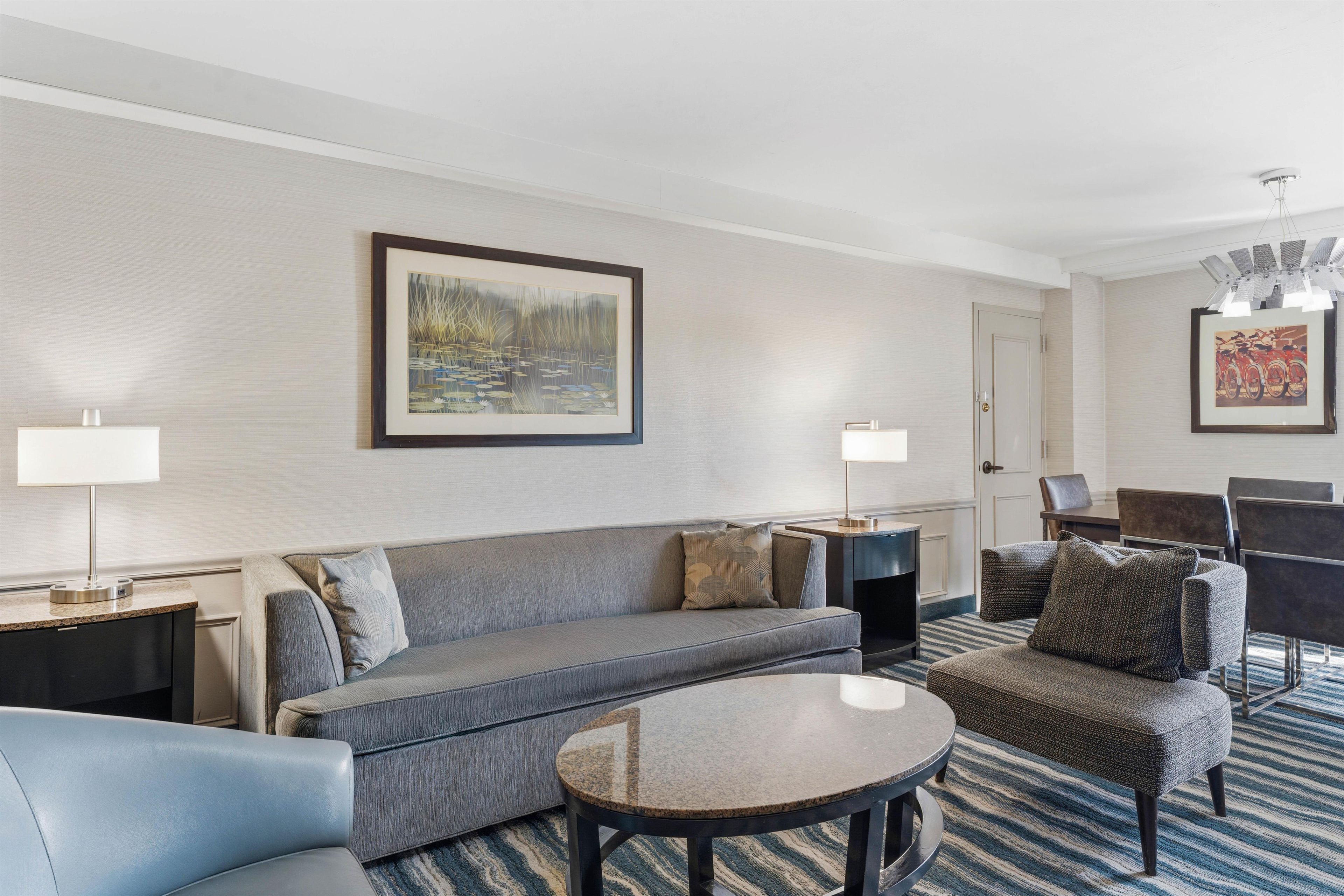 A modern hotel room with a grey sofa, a round coffee table, a patterned armchair, a dining area with a table and chairs, and a painting on the wall.