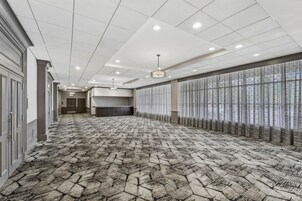 Un pasillo de hotel con alfombra estampada, una larga recepción y una fila de ventanas con cortinas translúcidas.