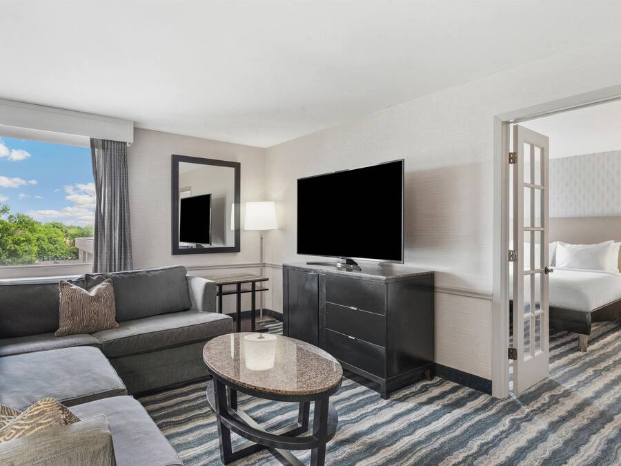 Una habitación de hotel con sofá, una mesita redonda, televisión, espejo, lámpara y cama.