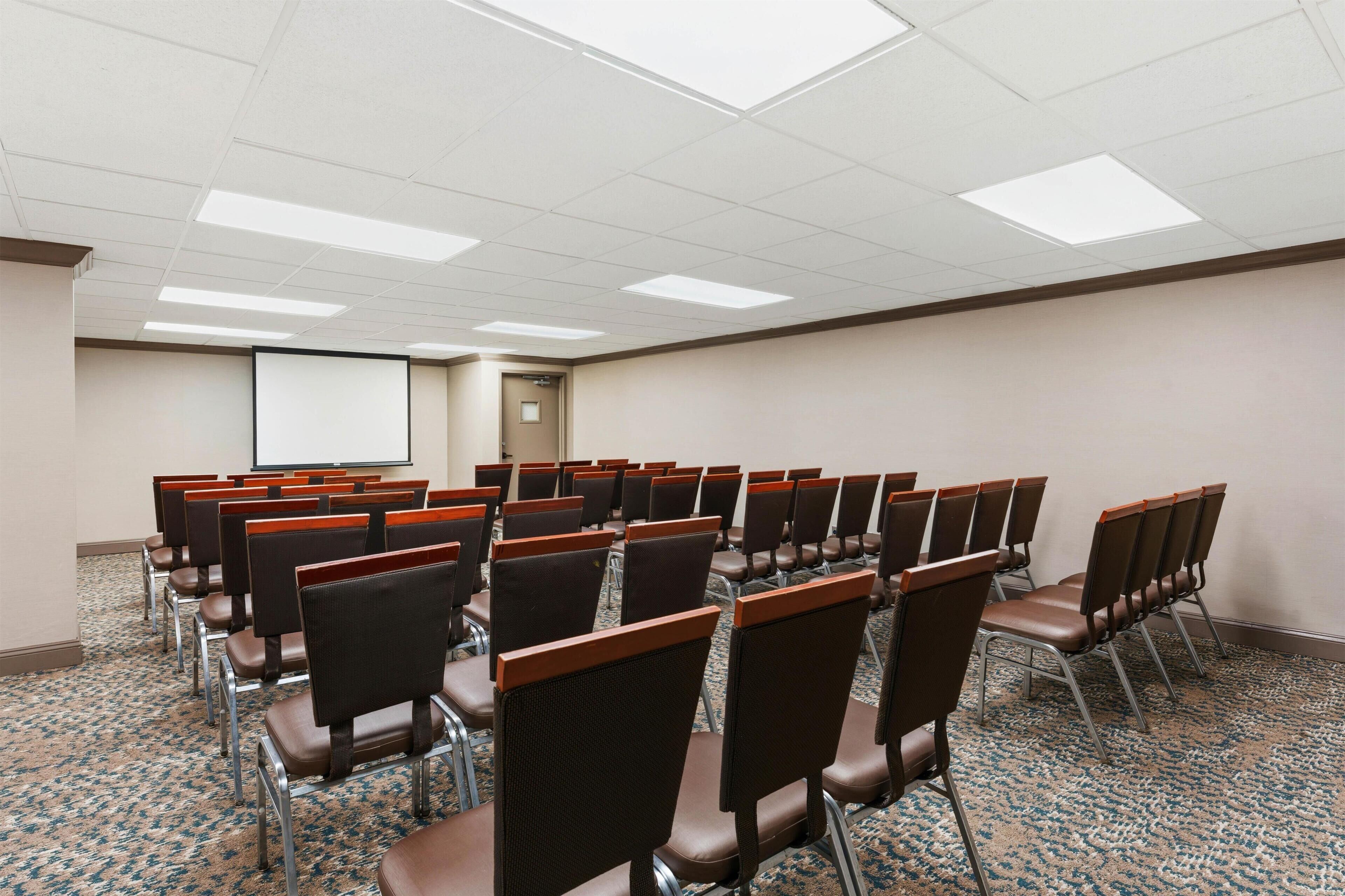 Un salón de conferencias con filas de sillas, una pantalla de proyector y piso alfombrado.