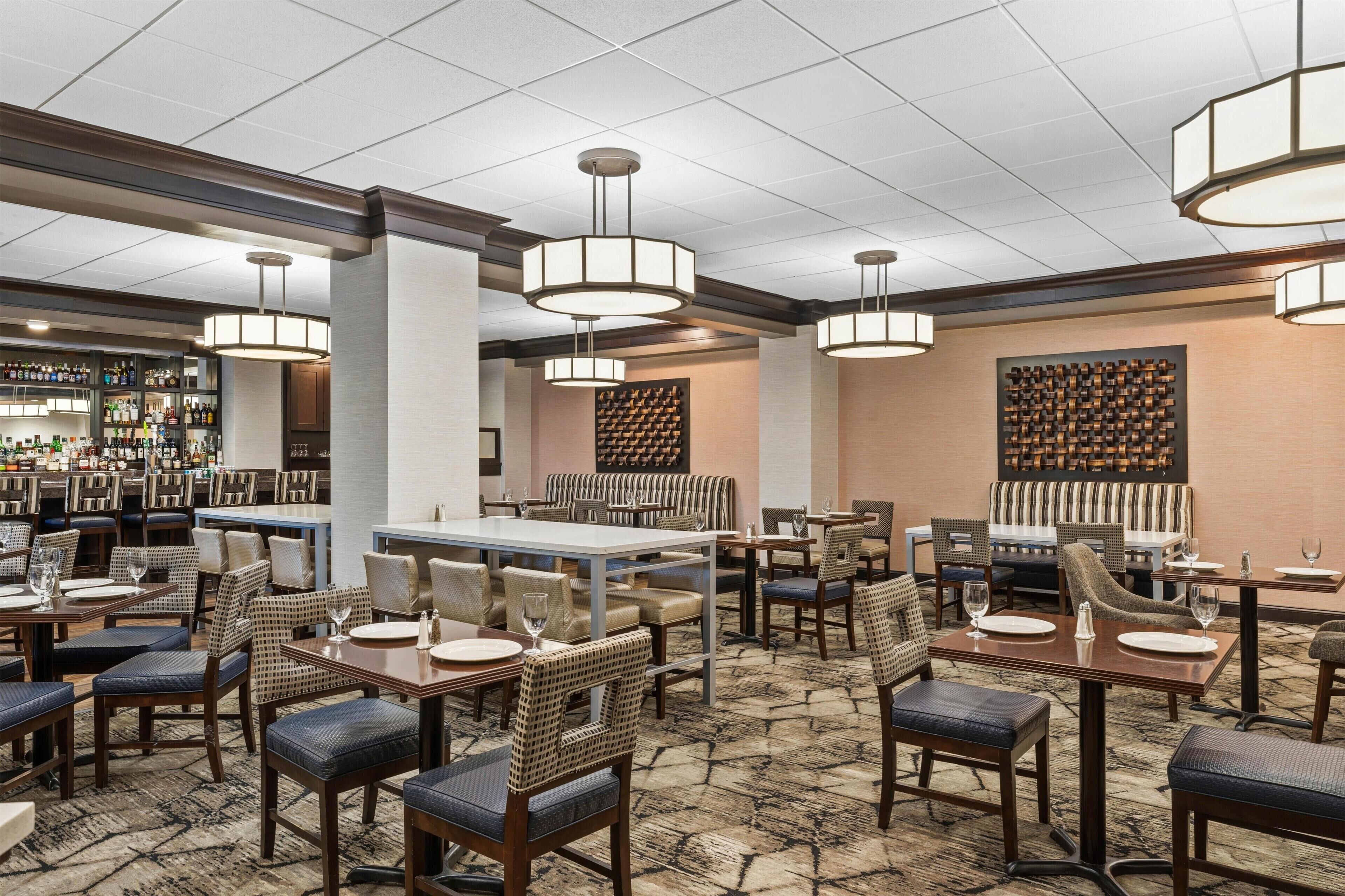Une salle à manger d’hôtel avec de nombreuses tables et chaises, un bar bien garni de différentes bouteilles et une moquette à motifs.