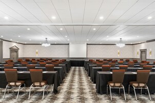 Un salón de conferencias con filas de mesas y sillas, piso alfombrado y techo blanco con iluminación empotrada.