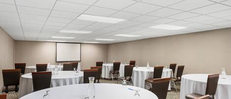 A conference room with multiple round tables, chairs, and a projector screen on the wall.