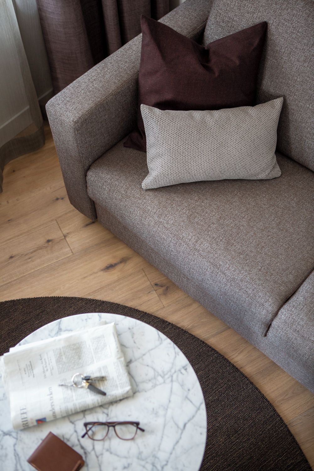 A grey sofa with a patterned pillow, a round coffee table with a newspaper, glasses, and a wallet.