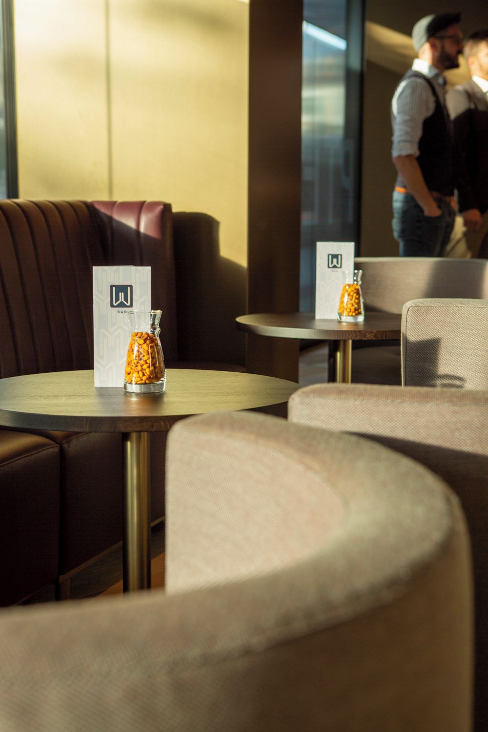 A modern interior with round tables and cushioned seating, a glass jar with a label on one table, and a partial view of a person in the background.