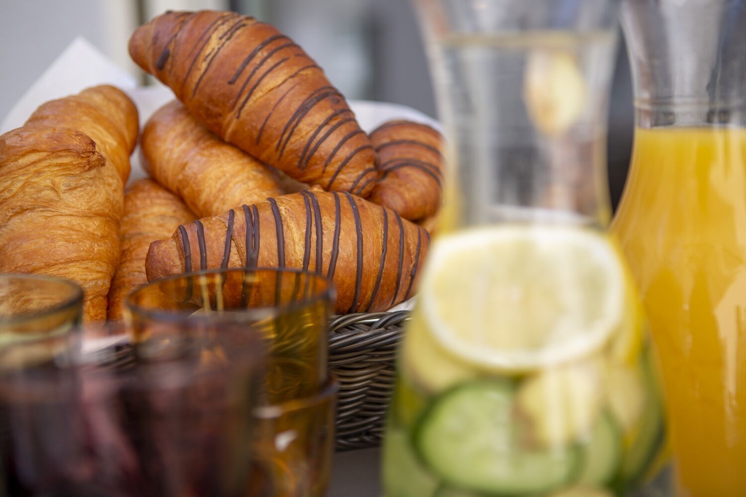 A breakfast setting with croissants, a basket, a glass of orange juice, and a glass with a slice of lemon.