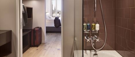 A modern bathroom with a bathtub, shower, and toiletries. There is a suitcase and a bed visible in the adjacent room.