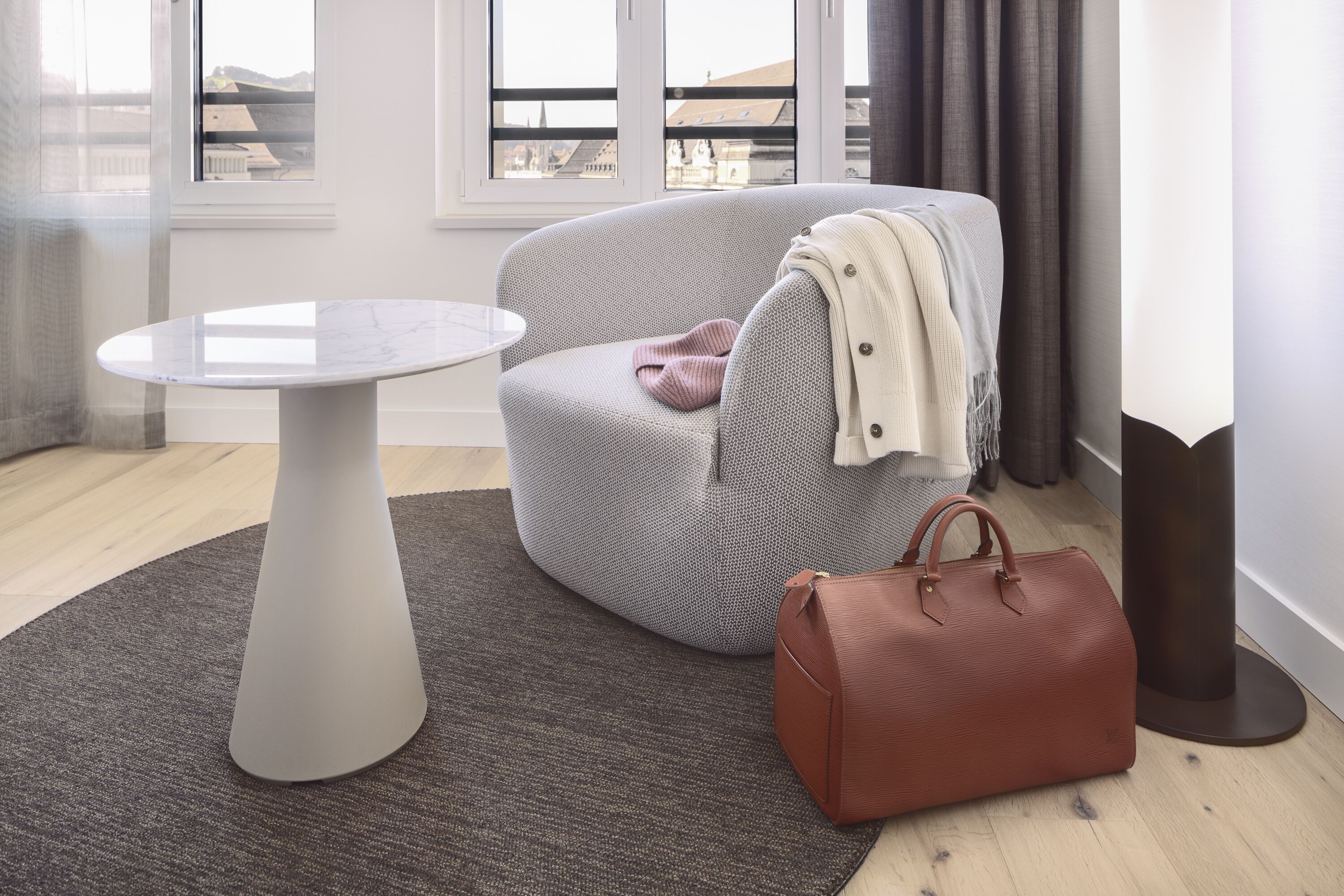 Une pièce moderne avec une table ronde blanche, un fauteuil gris agrémenté d’une couverture et un sac à main marron posé sur un tapis gris.