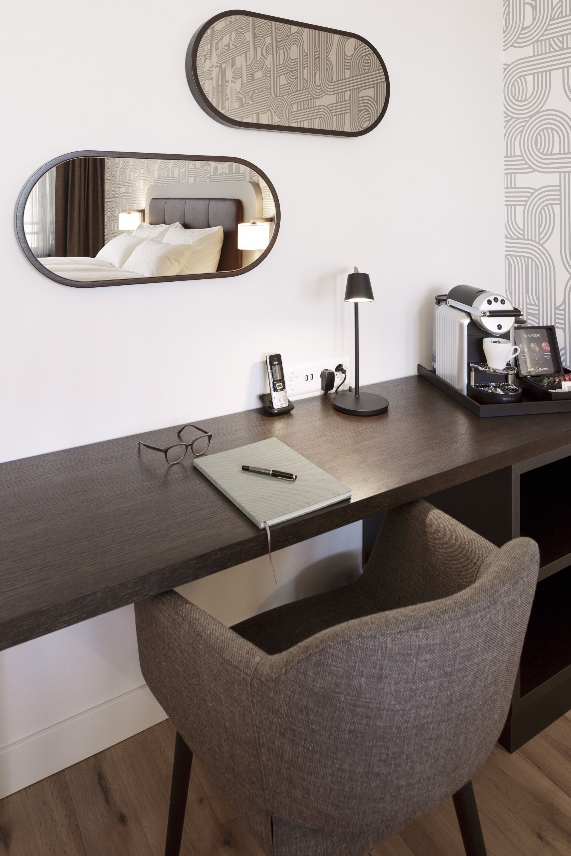Une chambre d’hôtel moderne avec un bureau en bois, une chaise, un lit avec une tête de lit, deux miroirs muraux, une machine à café, un téléphone et une paire de lunettes.