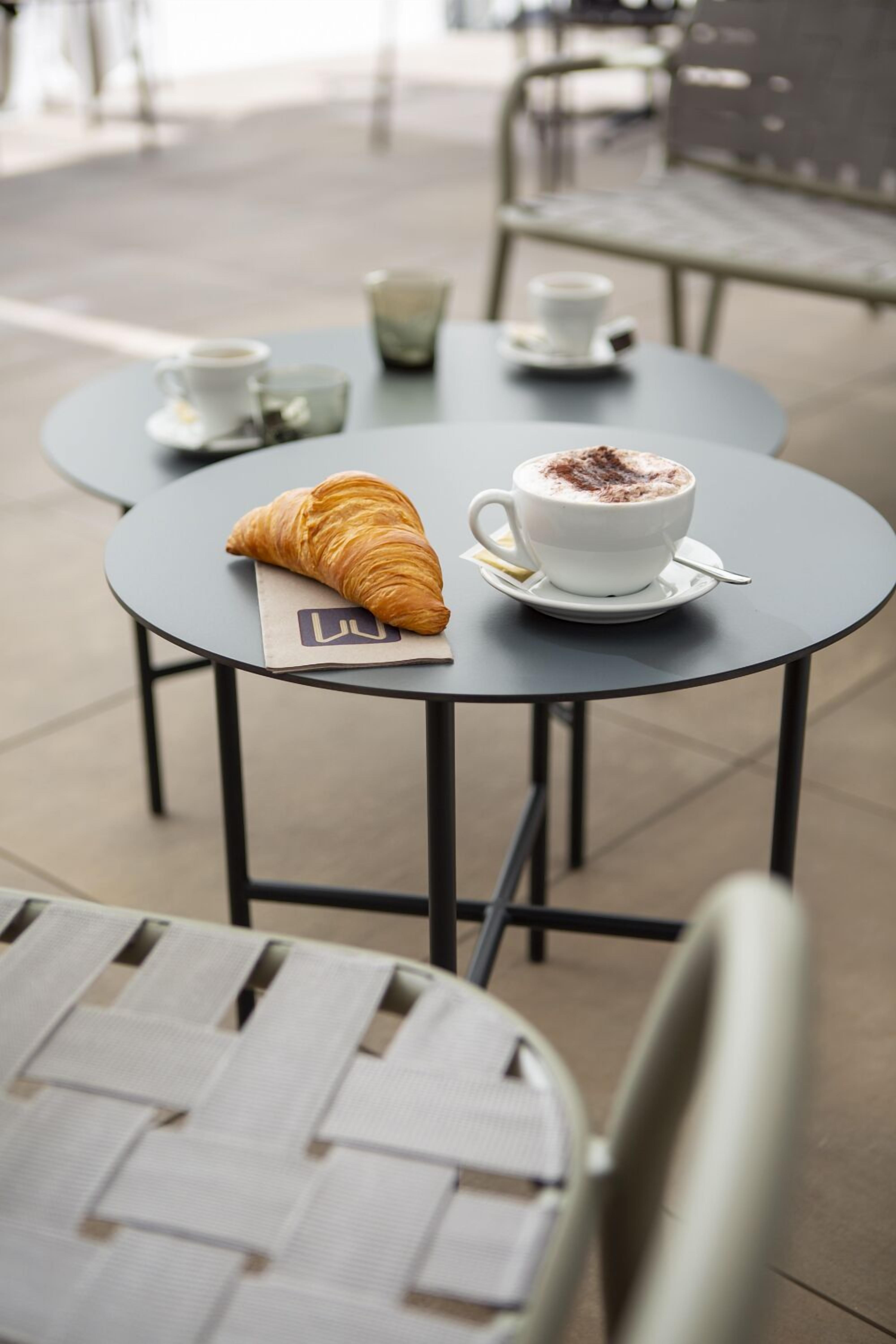 A round table with a croissant and a cup of coffee, another table with a cup of tea, and a chair in the foreground.