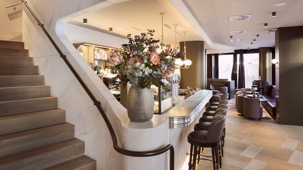 Un moderno vestíbulo de hotel con una escalera de mármol, una zona de bar con asientos y un jarrón grande con flores.