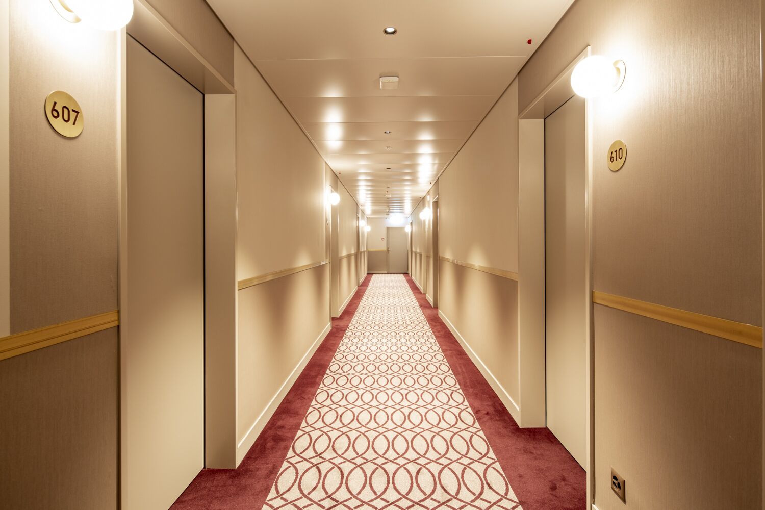 Pasillo de hotel con los números 607 y 610 en las paredes, una alfombra estampada y una moqueta roja que conduce al final.