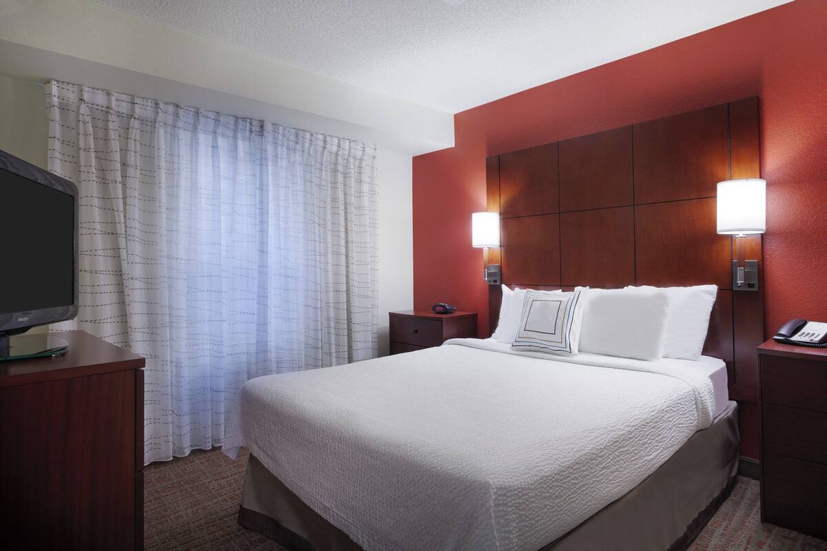 A hotel room with a large bed, a flat-screen TV on a wooden dresser, a nightstand with a lamp, and a red accent wall.