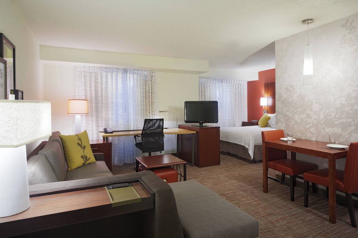 A hotel room with a sofa, a desk with a computer, a TV, a bed, and a dining table with chairs.