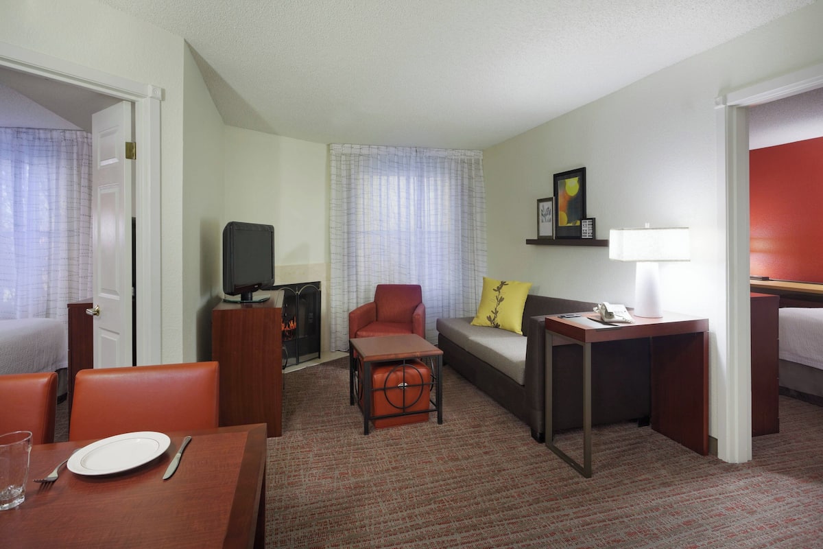 A hotel room with a dining table, a sofa, a small table with a lamp, a television, and a fireplace.