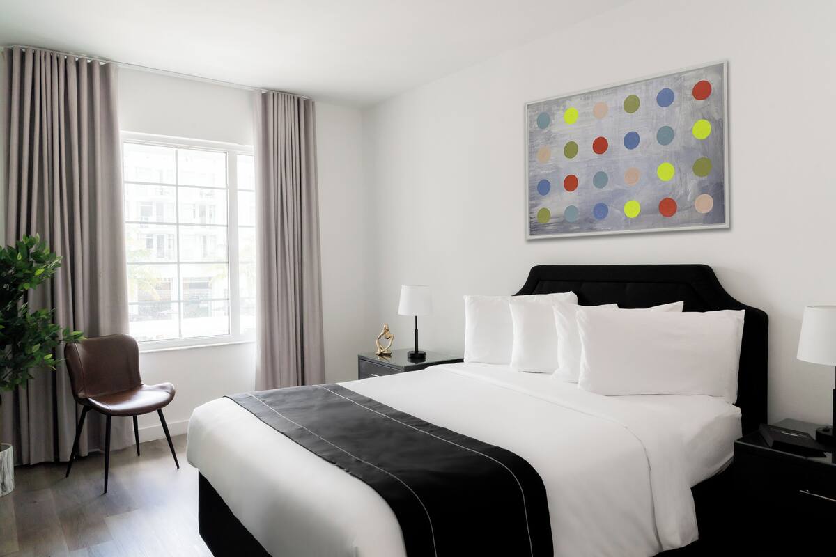 A modern hotel room with a large bed, a side table, a chair, and a window with curtains.