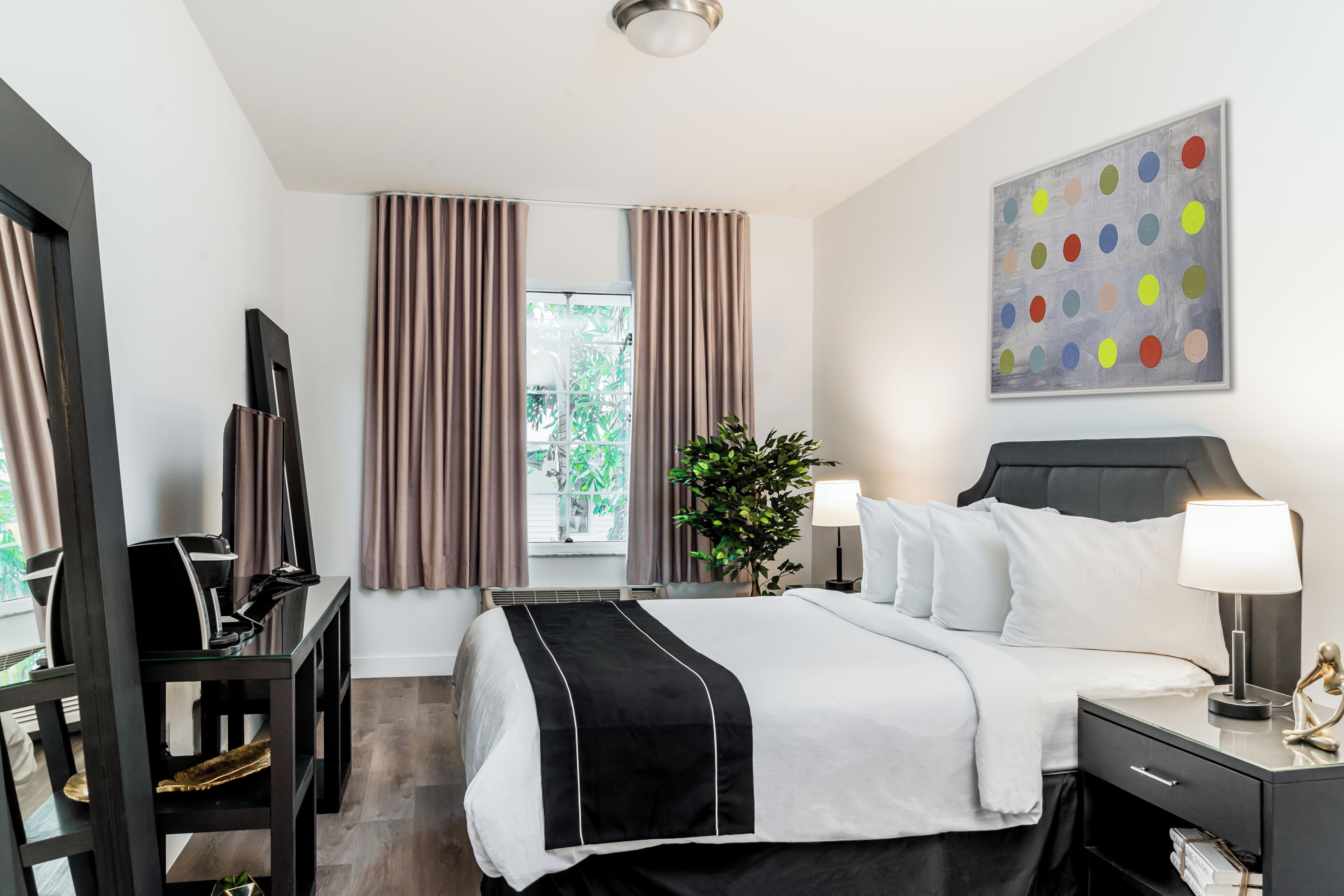 Une chambre d’hôtel comprenant un lit, des tables de chevet, un bureau, une chaise, un miroir, une fenêtre avec des rideaux, une plante et un tableau au mur.