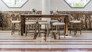 Comedor con mesa de madera y seis taburetes, ventanas octogonales y pared de mármol.