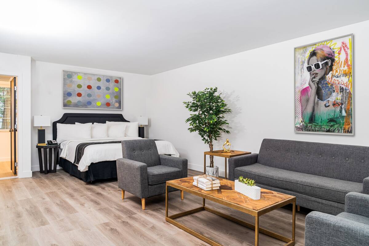 A modern hotel room with a bed, grey sofas, a wooden coffee table, and a large abstract painting.