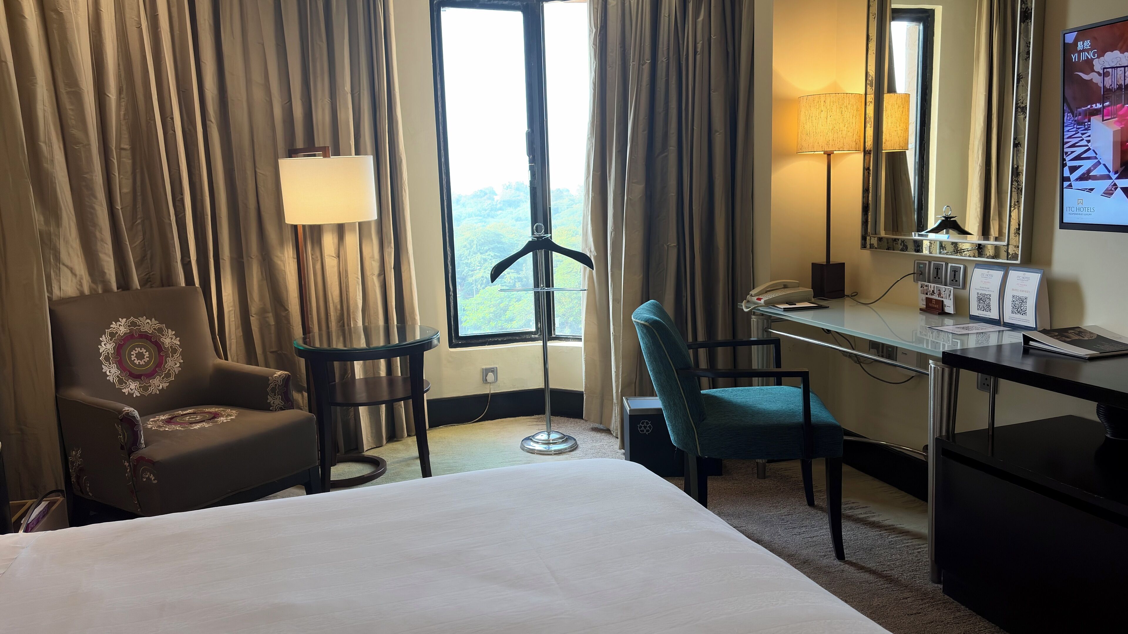 A hotel room with a bed, a desk, a chair, a lamp, and a window with curtains.