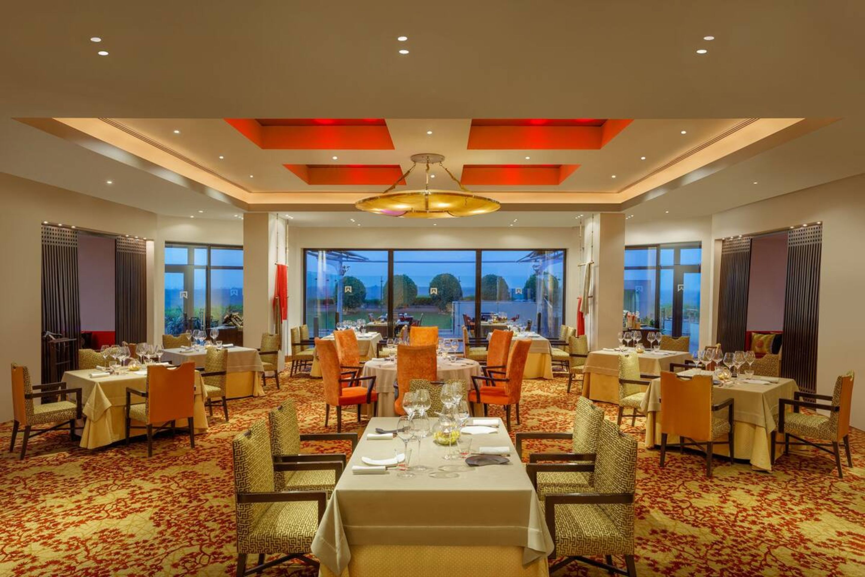 Un comedor de hotel con varias mesas dispuestas para los huéspedes, amplias ventanas que ofrecen vistas al exterior y un diseño de techo moderno.