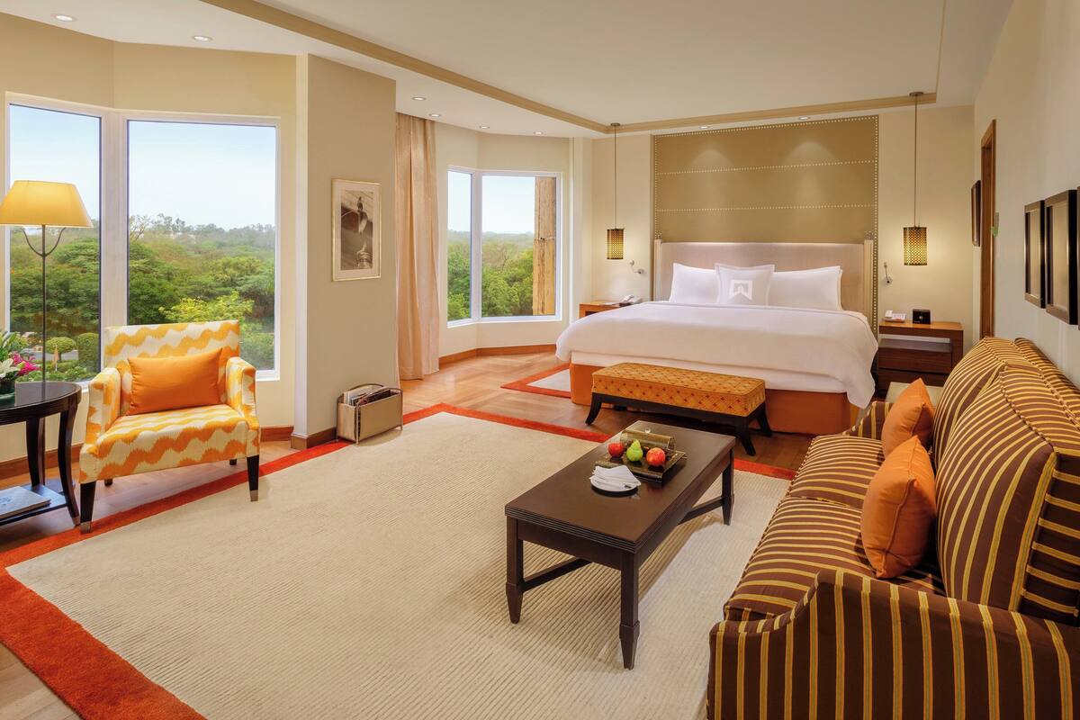 A spacious hotel room with a large bed, a sofa, a coffee table, and a view of greenery through the windows.