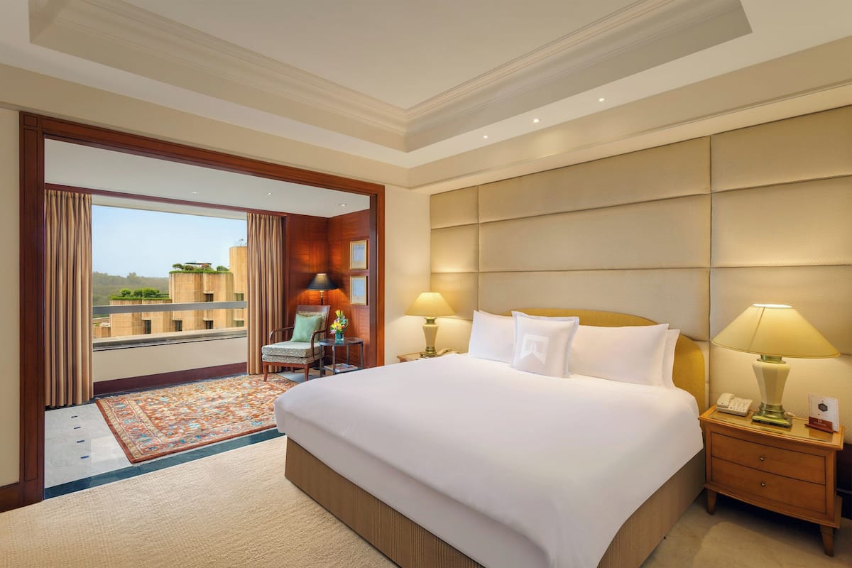 A hotel room with a large bed, two bedside lamps, a nightstand, a chair, a small table, and a view of the outside.