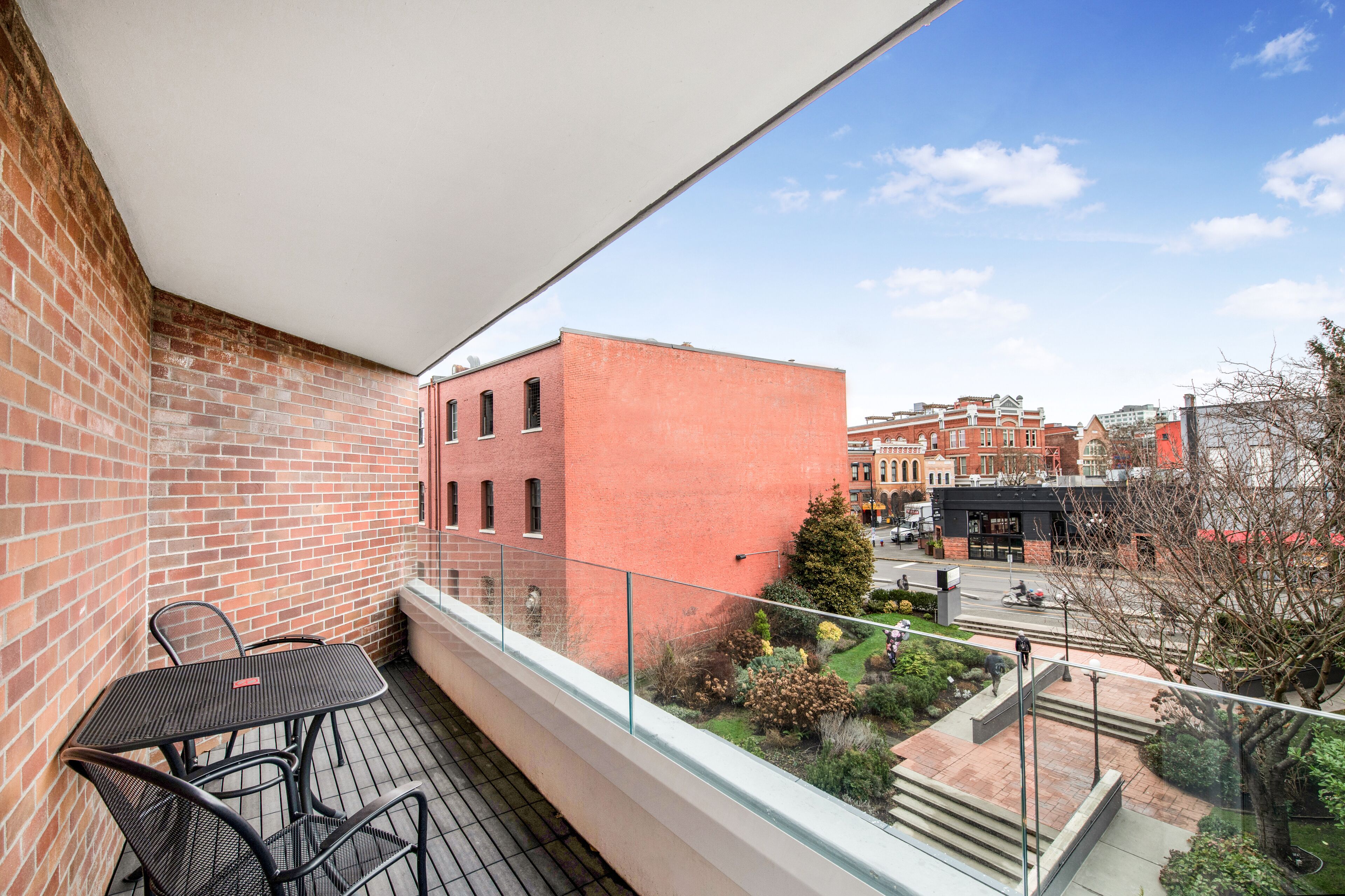 A balcony with a brick wall, a glass railing, and a view of a cityscape with buildings and a tree.