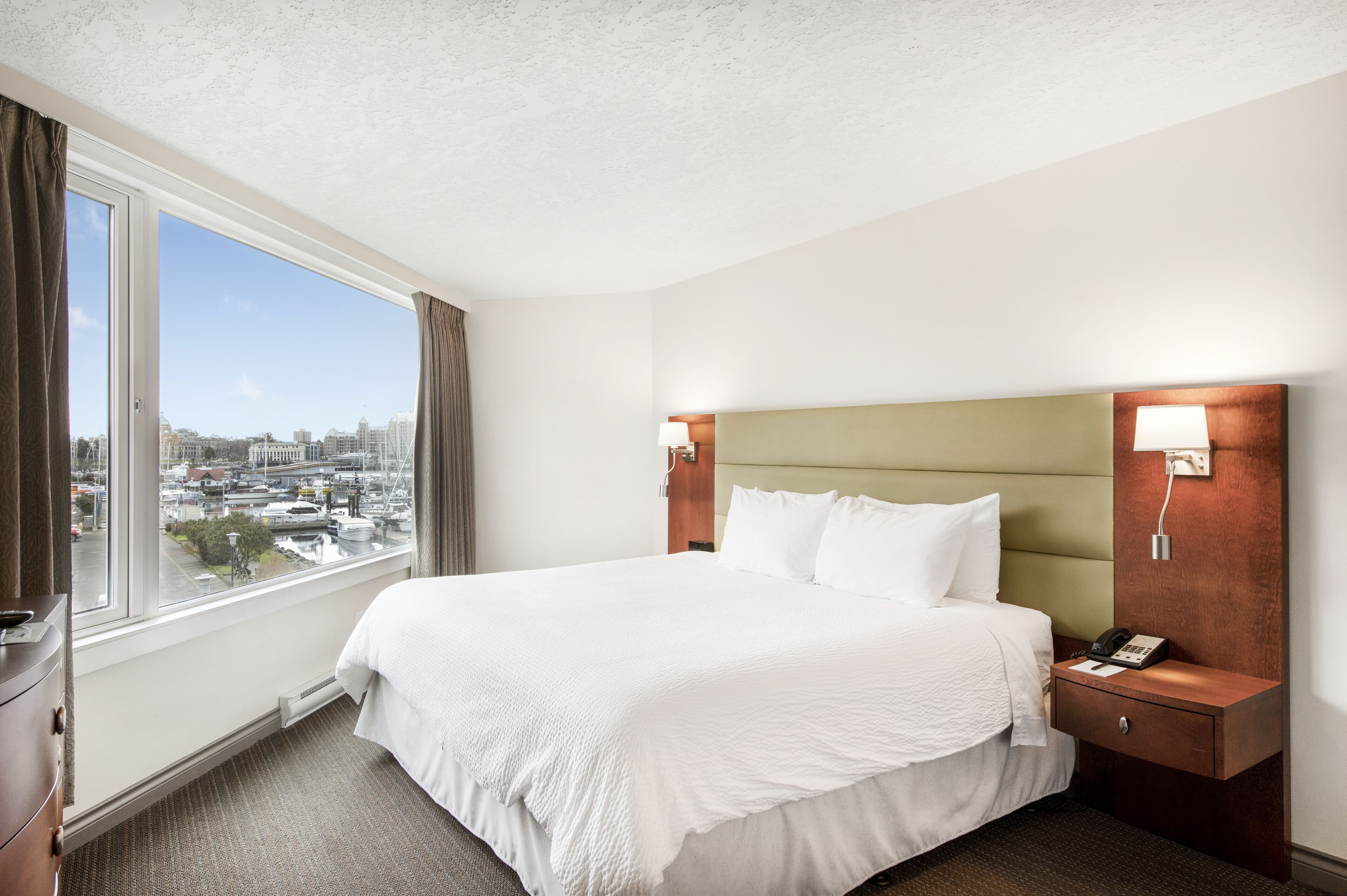 Habitación de hotel con una cama grande, una mesita de noche, una lámpara y vistas a un paisaje urbano a través de un ventanal amplio.