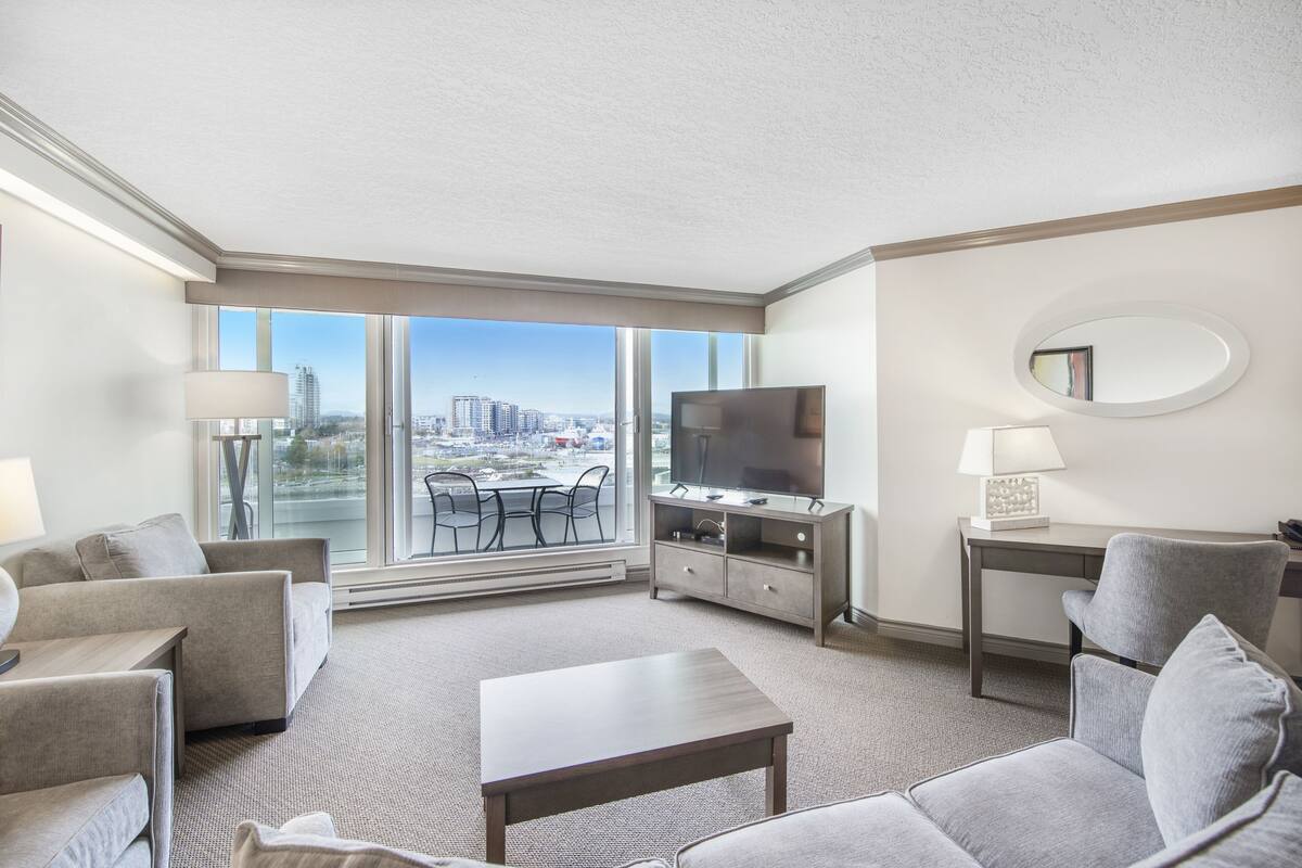 Ein modernes Hotelzimmer mit großem Fenster und Blick auf die Stadt, einem Flachbildfernseher, einem Essbereich mit Tisch und Stühlen sowie einem kleinen Schreibtisch mit Lampe.