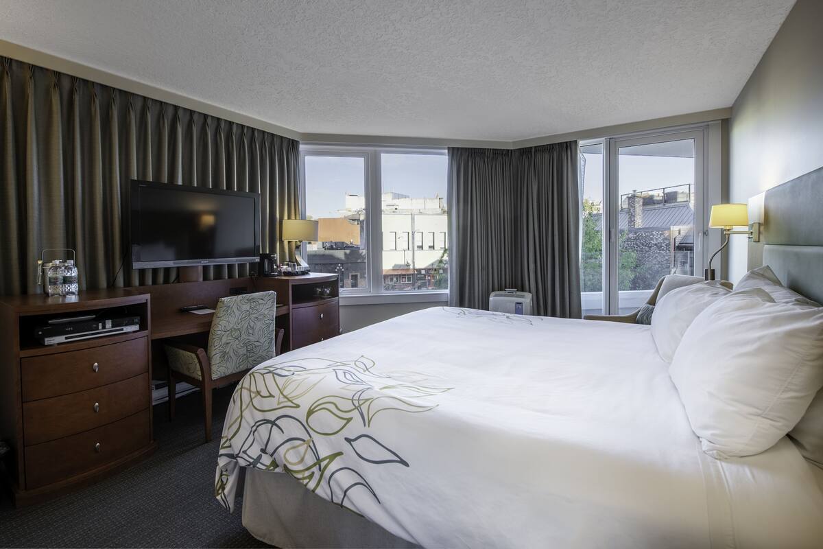 Ein Hotelzimmer mit einem großen Bett, einem Schreibtisch mit Fernseher, einem Sessel und Blick auf Gebäude durch das Fenster.