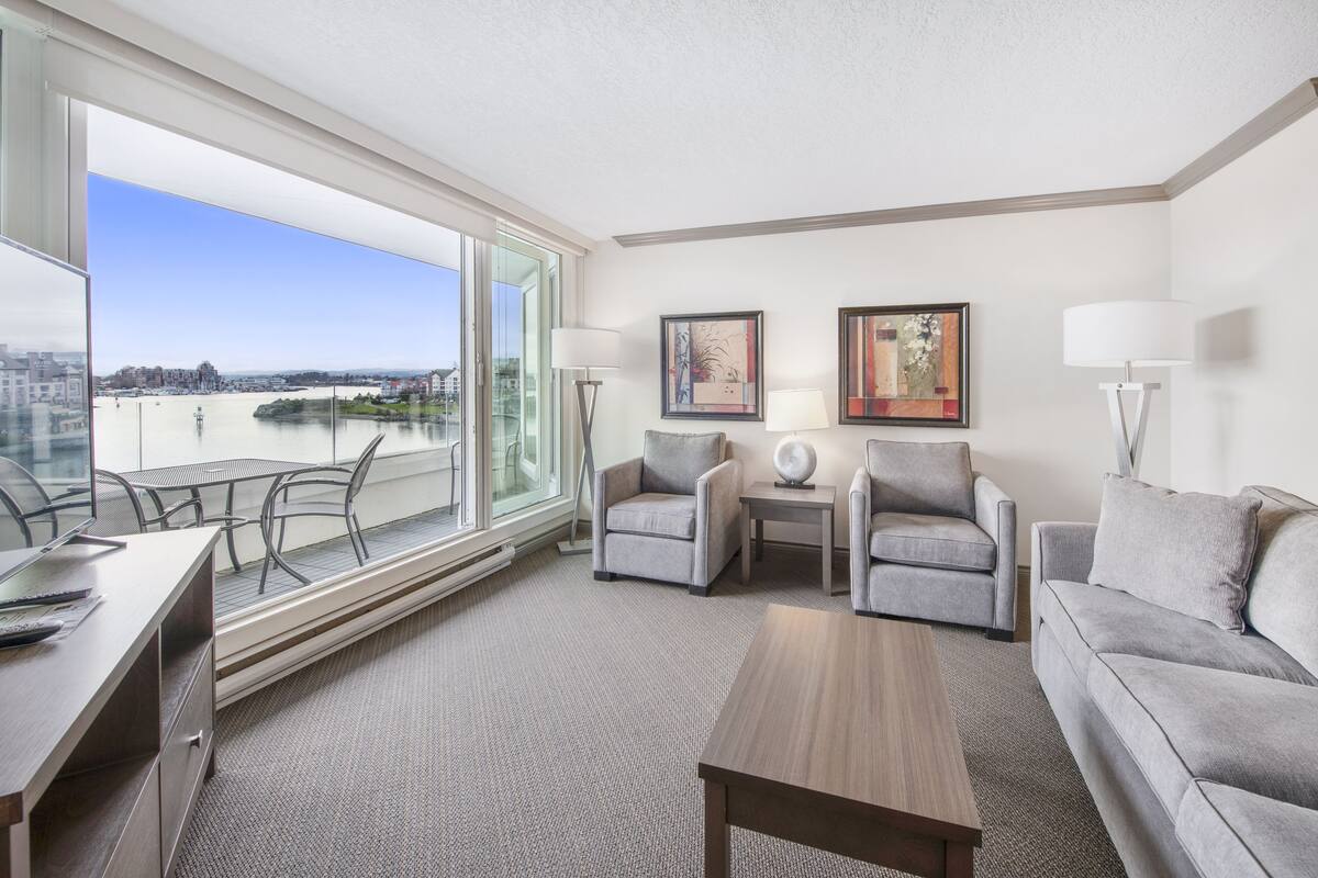 Ein modernes Hotelzimmer mit großem Fenster und Blick auf die Stadt, einem Schreibtisch mit Stuhl, einer Couch, zwei Sesseln, einem Couchtisch und zwei Wandleuchten.