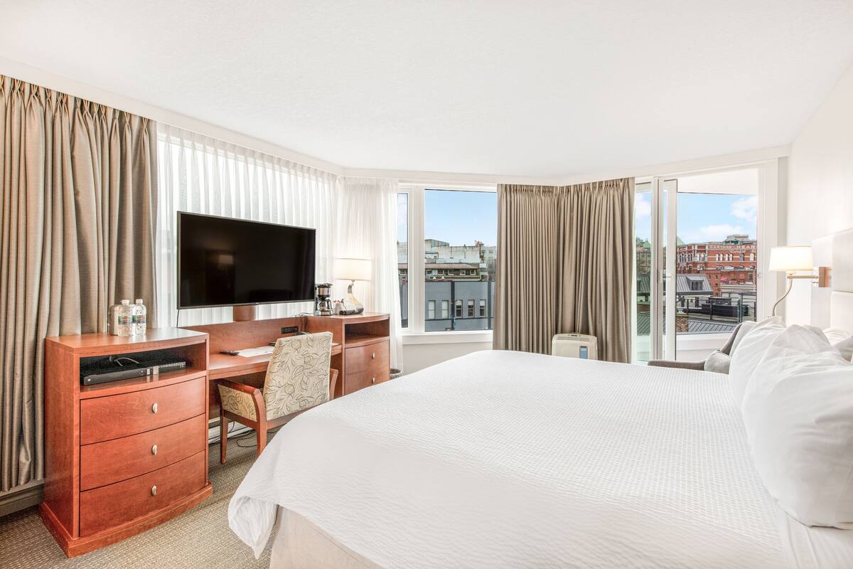 Ein Hotelzimmer mit einem großen Bett, einem Schreibtisch mit Flachbildfernseher, einem Sessel und einem Fenster mit Vorhängen.