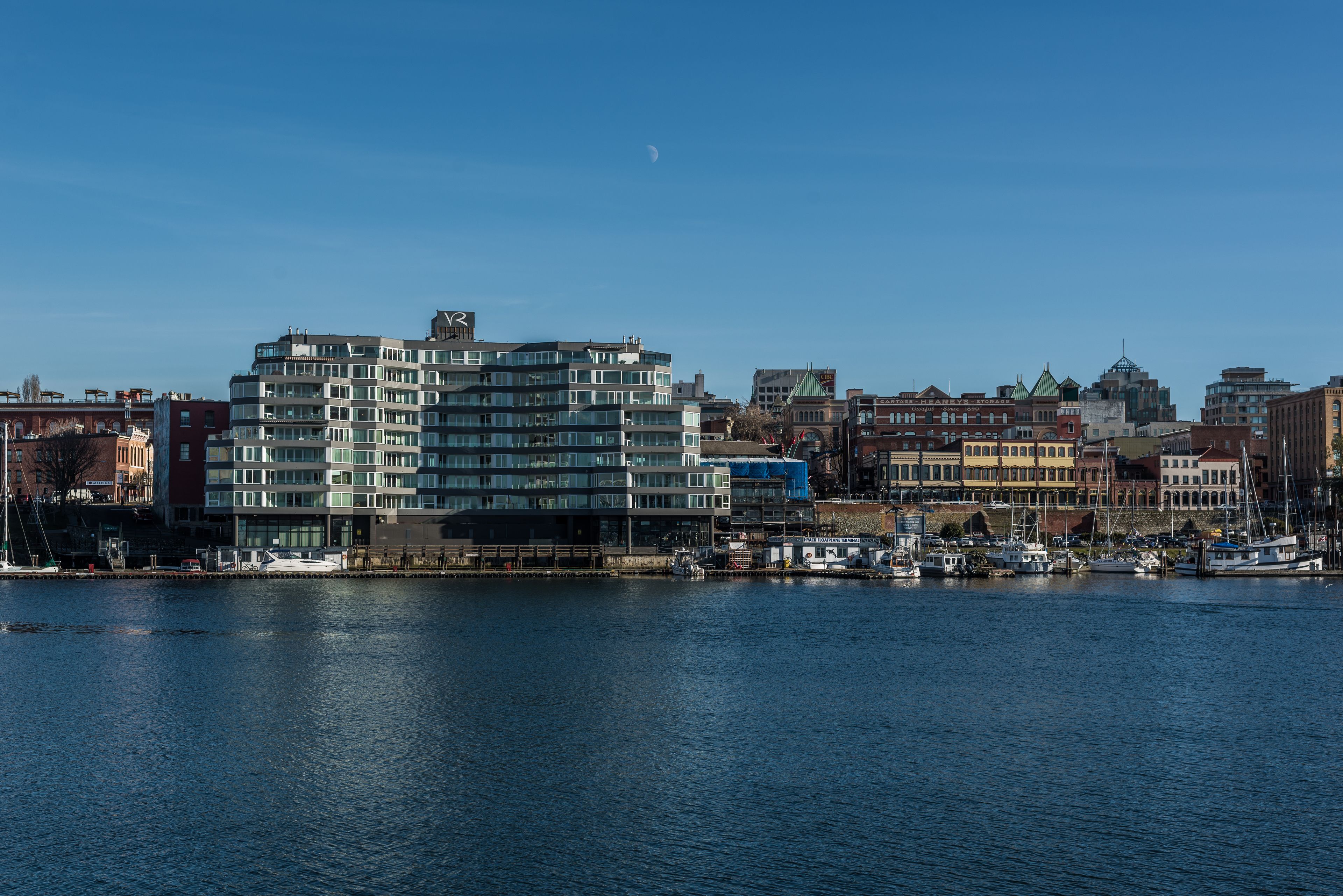 A waterfront cityscape with modern and historic buildings, a marina with boats, and a clear sky.