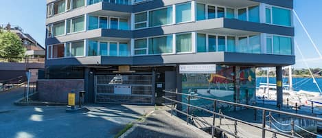 Un edificio moderno de varios pisos con balcones, una entrada porticada y vista a barcos en el fondo.
