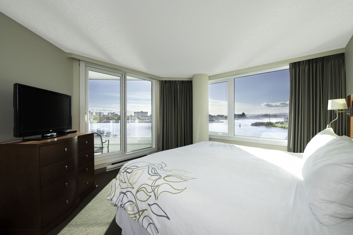 A hotel room with a large bed, a flat-screen TV on a dresser, and a view of a body of water through large windows.