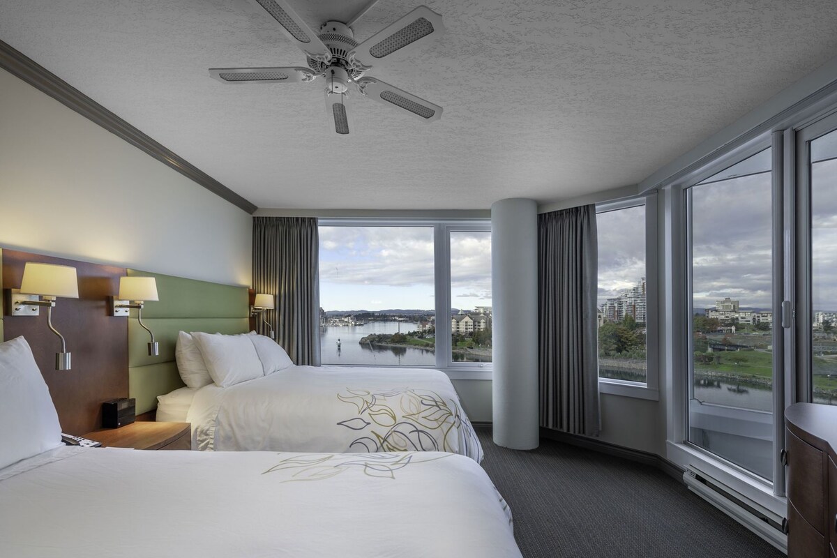 Ein Hotelzimmer mit einem großen Bett, einem Schreibtisch, einem Sessel, einem Deckenventilator und Blick auf einen Fluss sowie Gebäude.