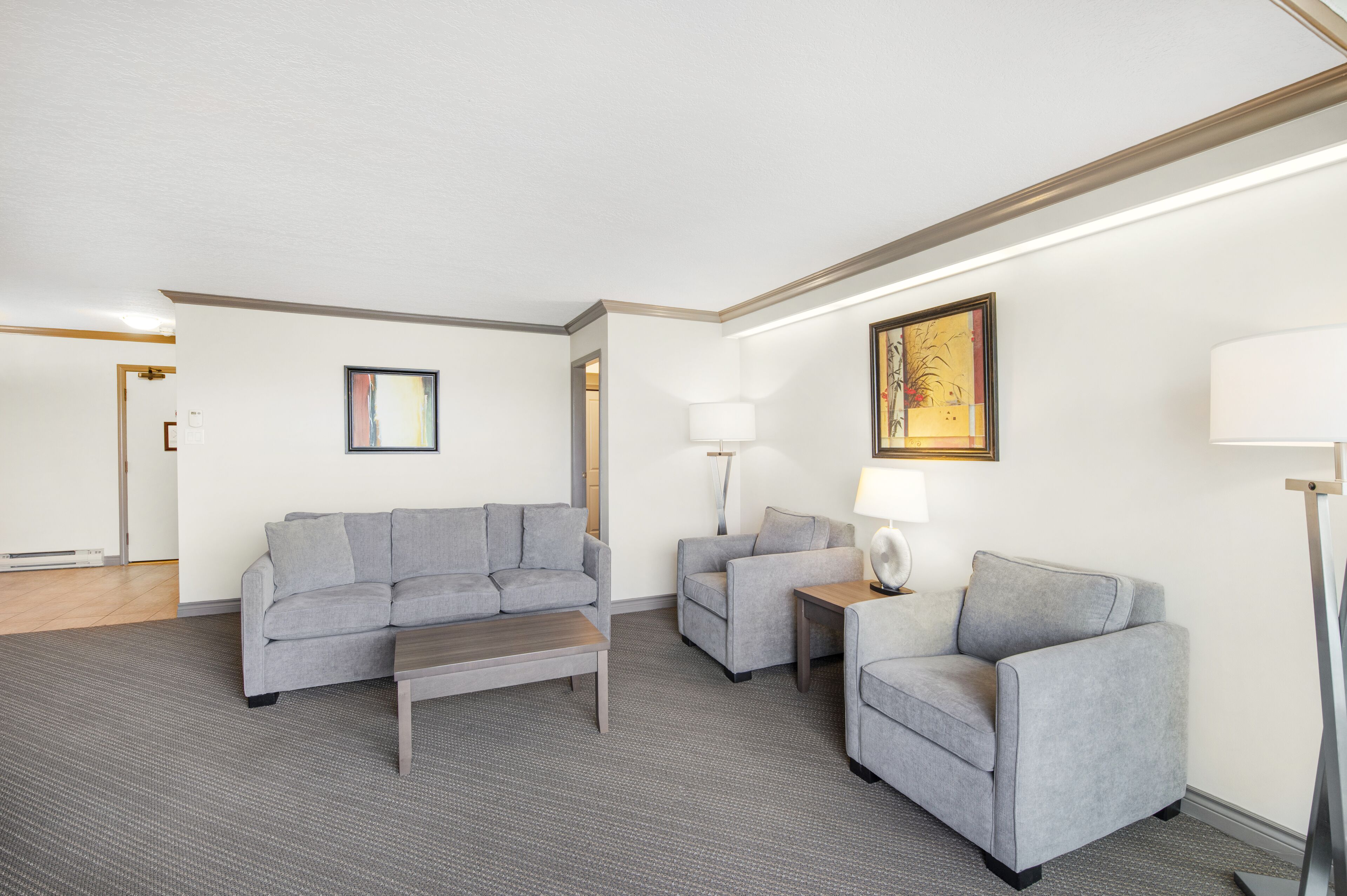 Un salon moderne avec un canapé gris, une table basse et deux fauteuils. On trouve deux lampes sur des tables d’appoint et un tableau au mur.