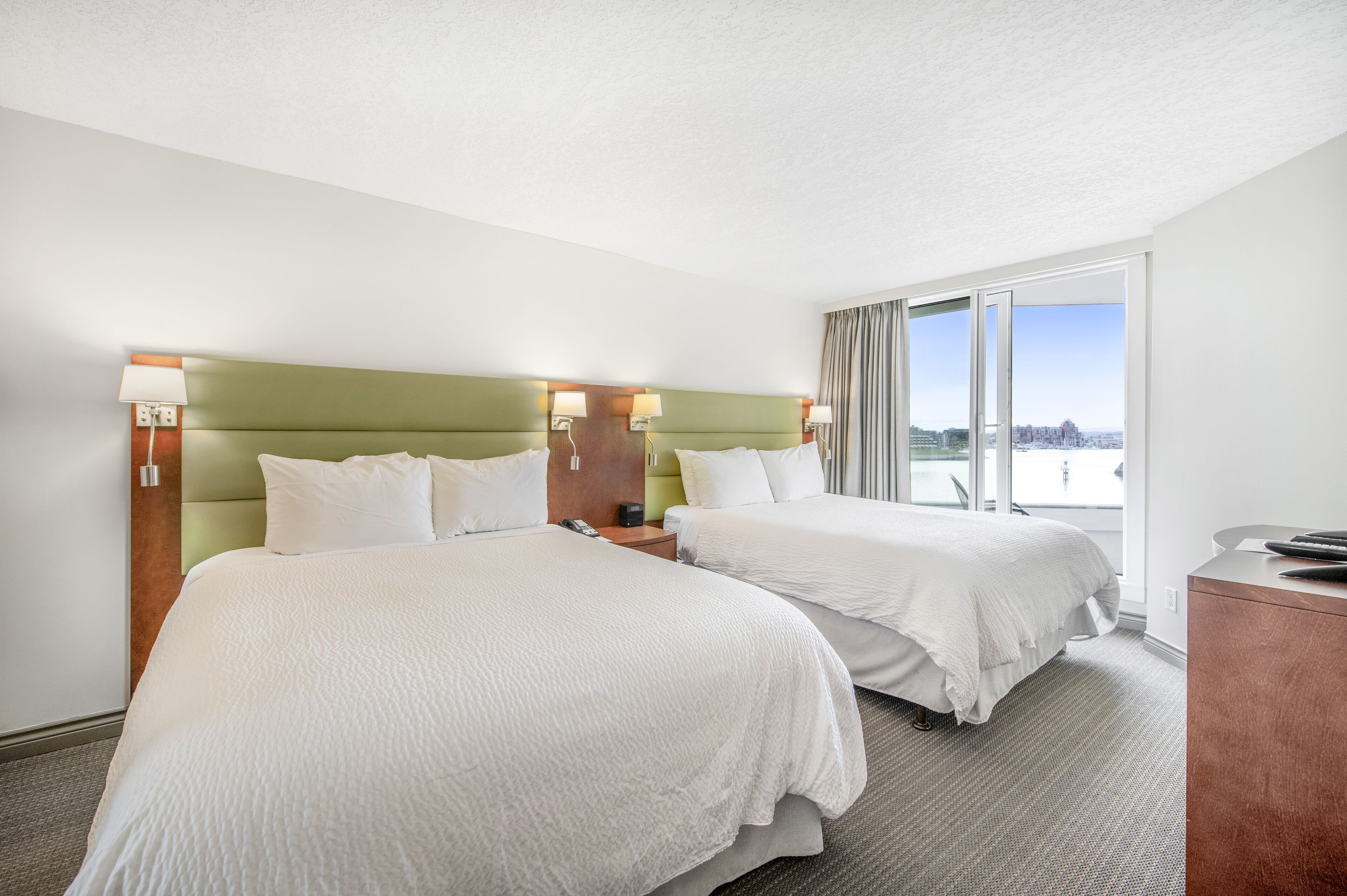 Hotelkamer met twee bedden, een groen hoofdeinde, een bureau en een groot raam met uitzicht op de stad.