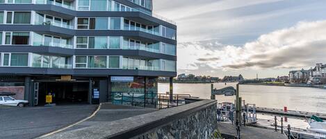 Um edifício moderno com vários andares e janelas amplas, situado perto de uma orla com um cais e barcos.