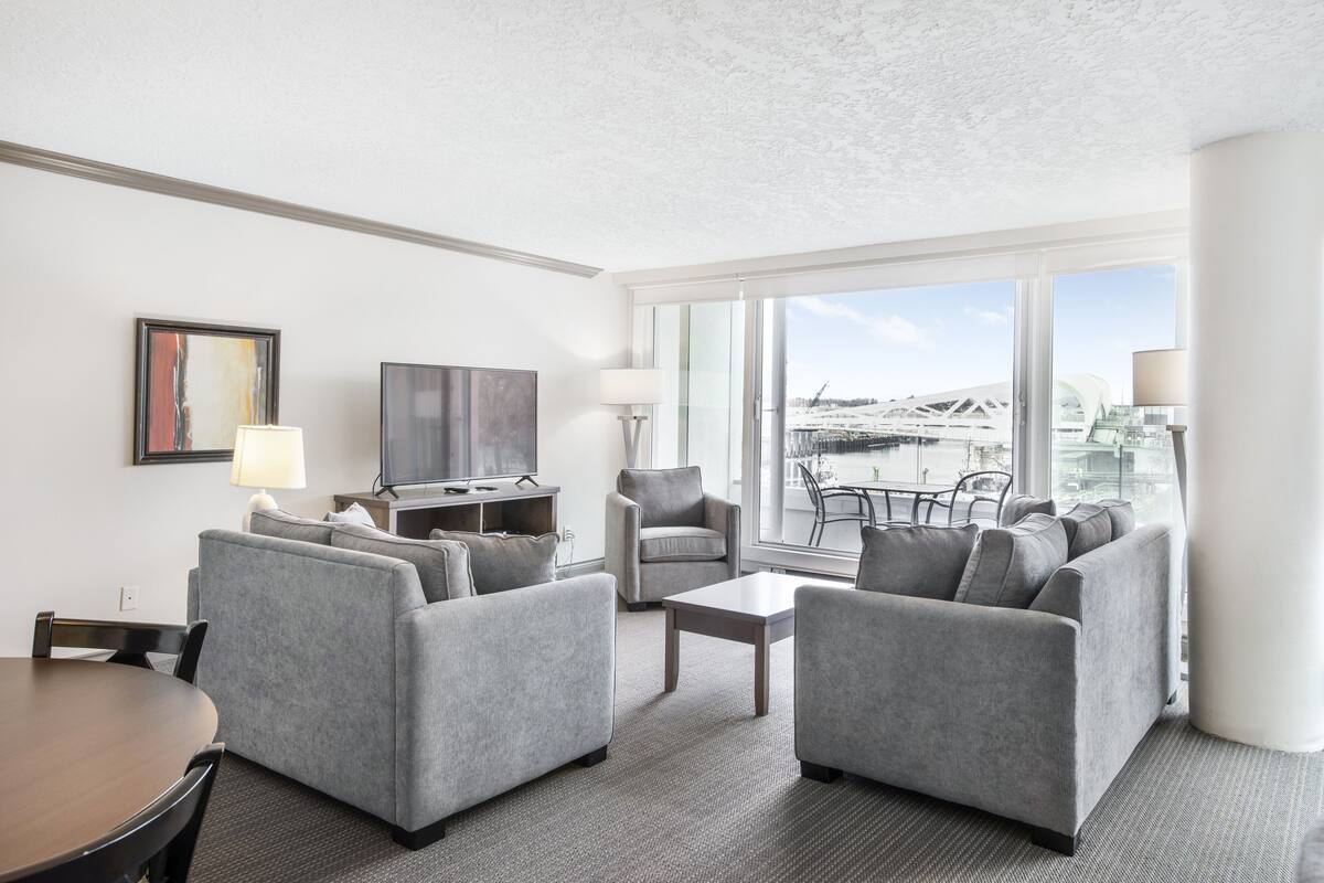 Ein modernes Hotelzimmer mit grauen Sofas, einem Esstisch, einem Flachbildfernseher und großen Fenstern mit Blick auf eine Stadtlandschaft.