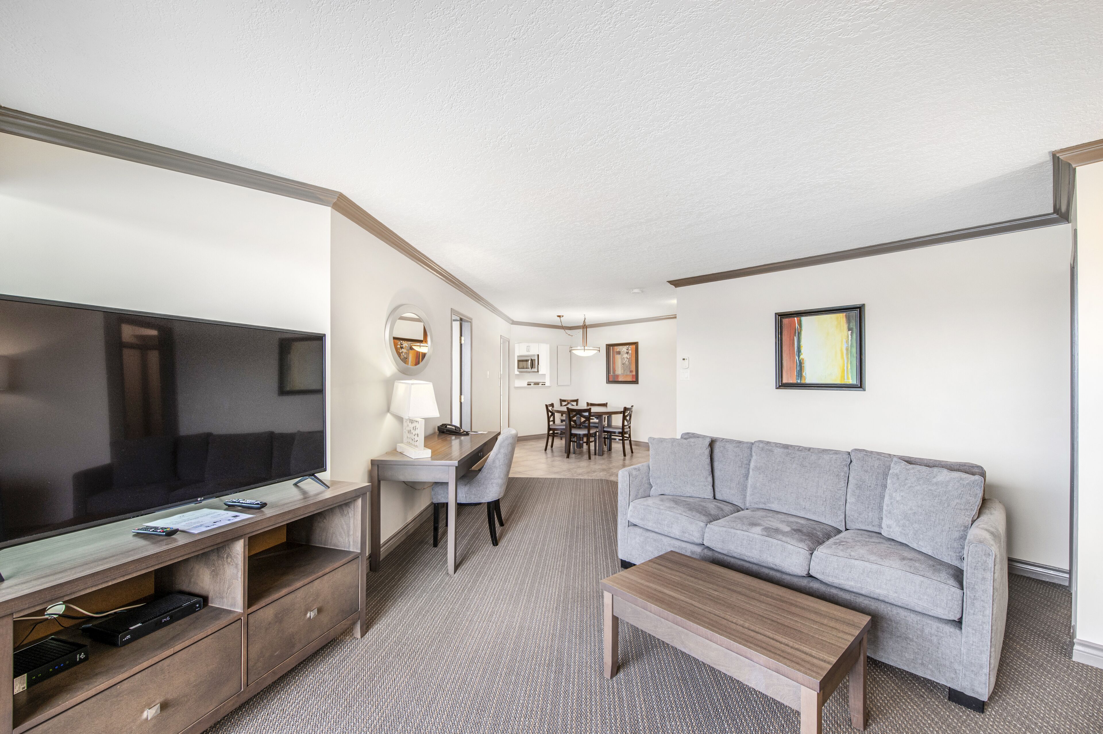 A modern hotel room with a flat-screen TV, a grey sofa, a wooden coffee table, a desk with a lamp, and a dining area in the background.
