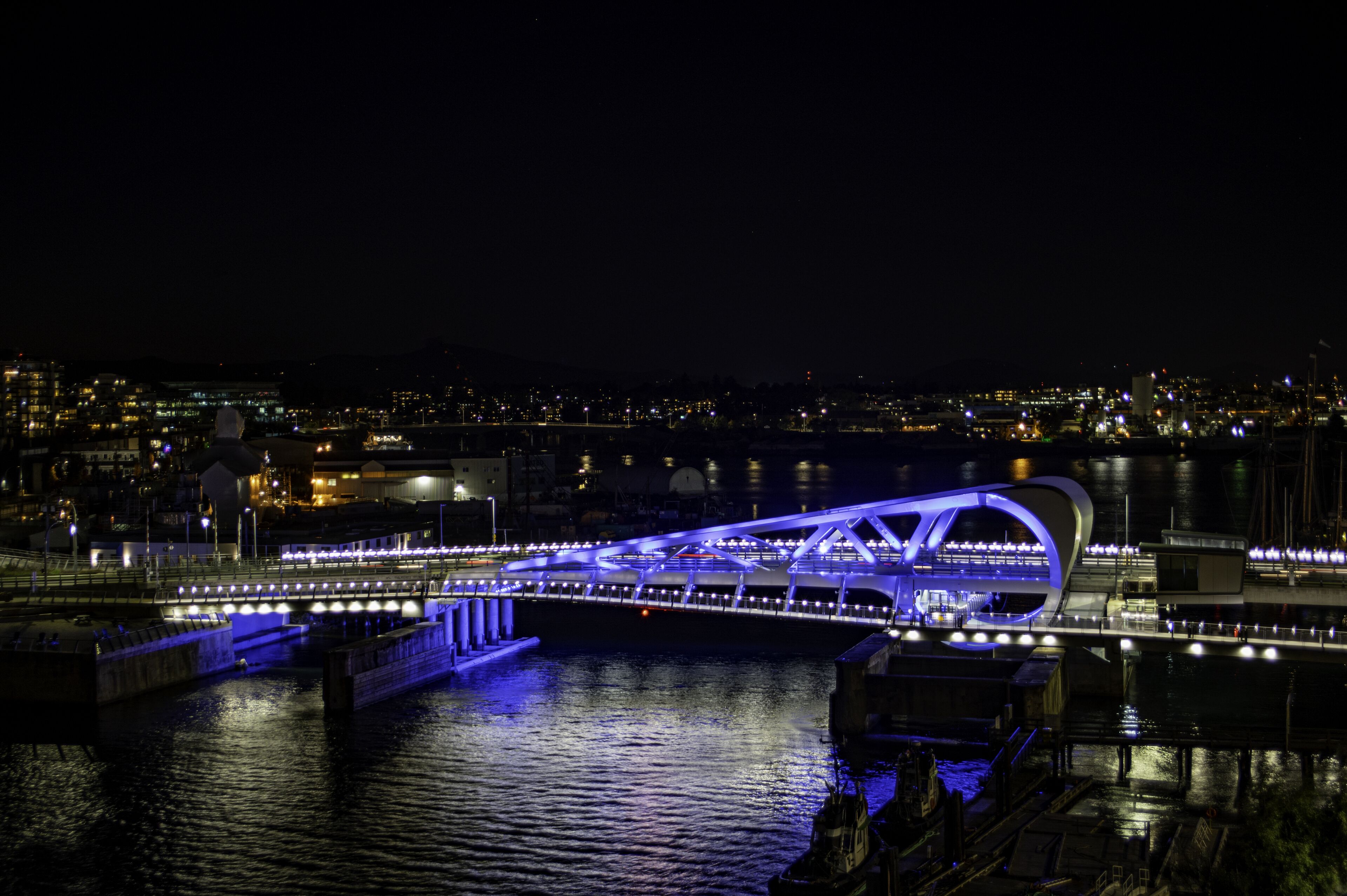 Une vue nocturne d’un pont moderne, illuminé de lumières bleues, qui enjambe une étendue d’eau, avec une vue sur la ville en arrière-plan.