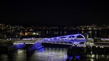 Une vue nocturne d’un pont moderne, illuminé de lumières bleues, qui enjambe une étendue d’eau, avec une vue sur la ville en arrière-plan.