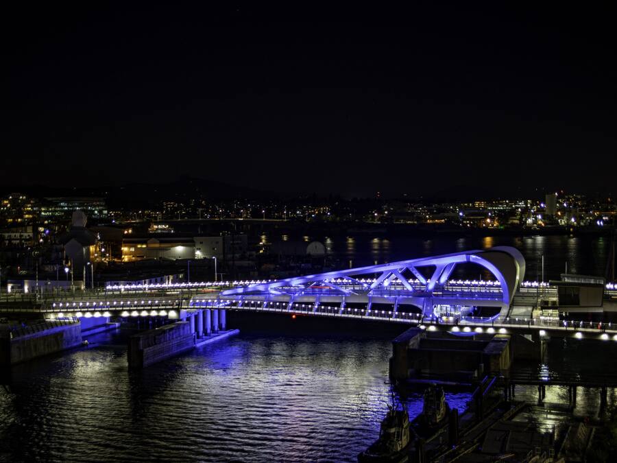 Une vue nocturne d’un pont moderne, illuminé de lumières bleues, qui enjambe une étendue d’eau, avec une vue sur la ville en arrière-plan.
