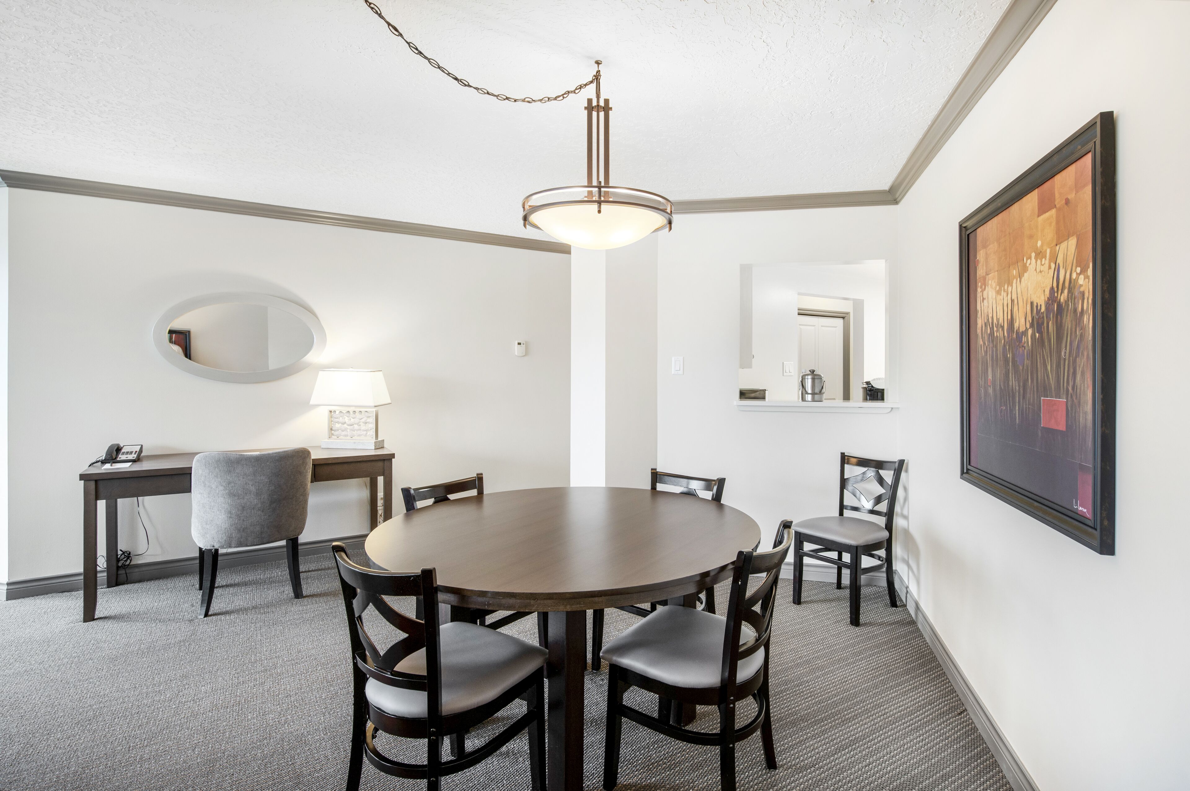 Habitación de hotel con una mesa redonda para comer, cuatro sillas, un escritorio con lámpara, un espejo y una obra de arte enmarcada en la pared.