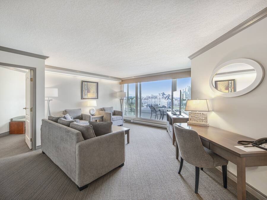 A hotel room with a sofa, a desk, a chair, a lamp, and a large window offering a city view.