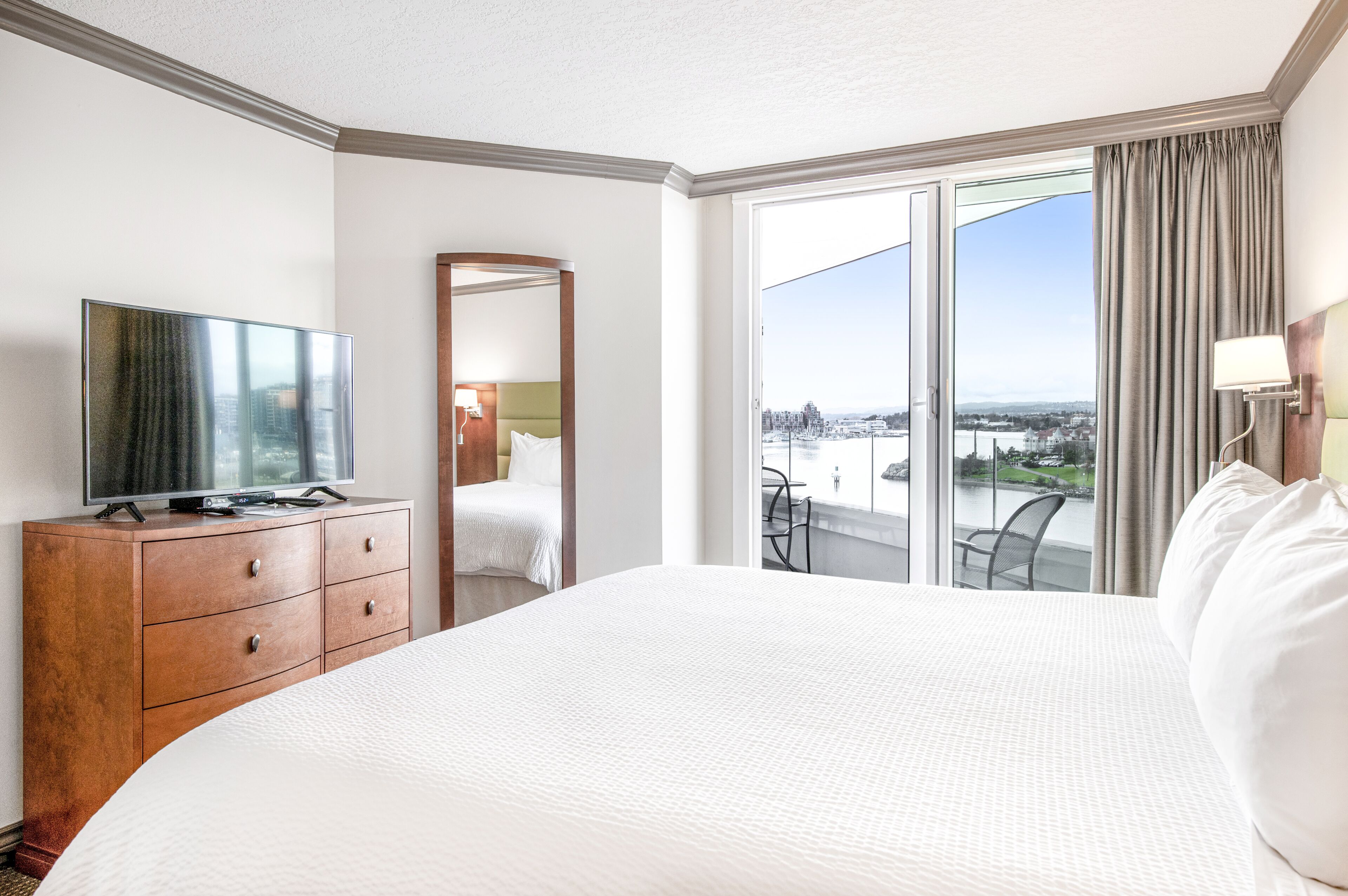 A hotel room with a large bed, a television on a wooden dresser, a balcony with a view of the city, and a sliding glass door.